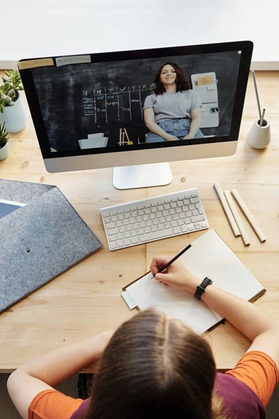 Top view of a student taking notes in a notebook during a live virtual classroom lecture on a desktop screen.