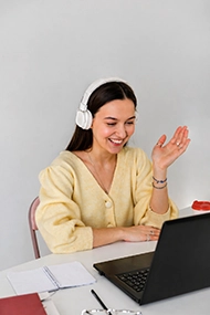 A smiling student wearing headphones and waving during an interactive online language learning class on a laptop.